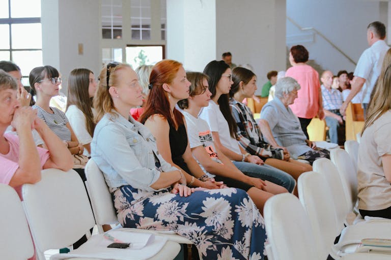 A diverse group of people sitting attentively during an indoor meeting or event, captured in a spacious room.
