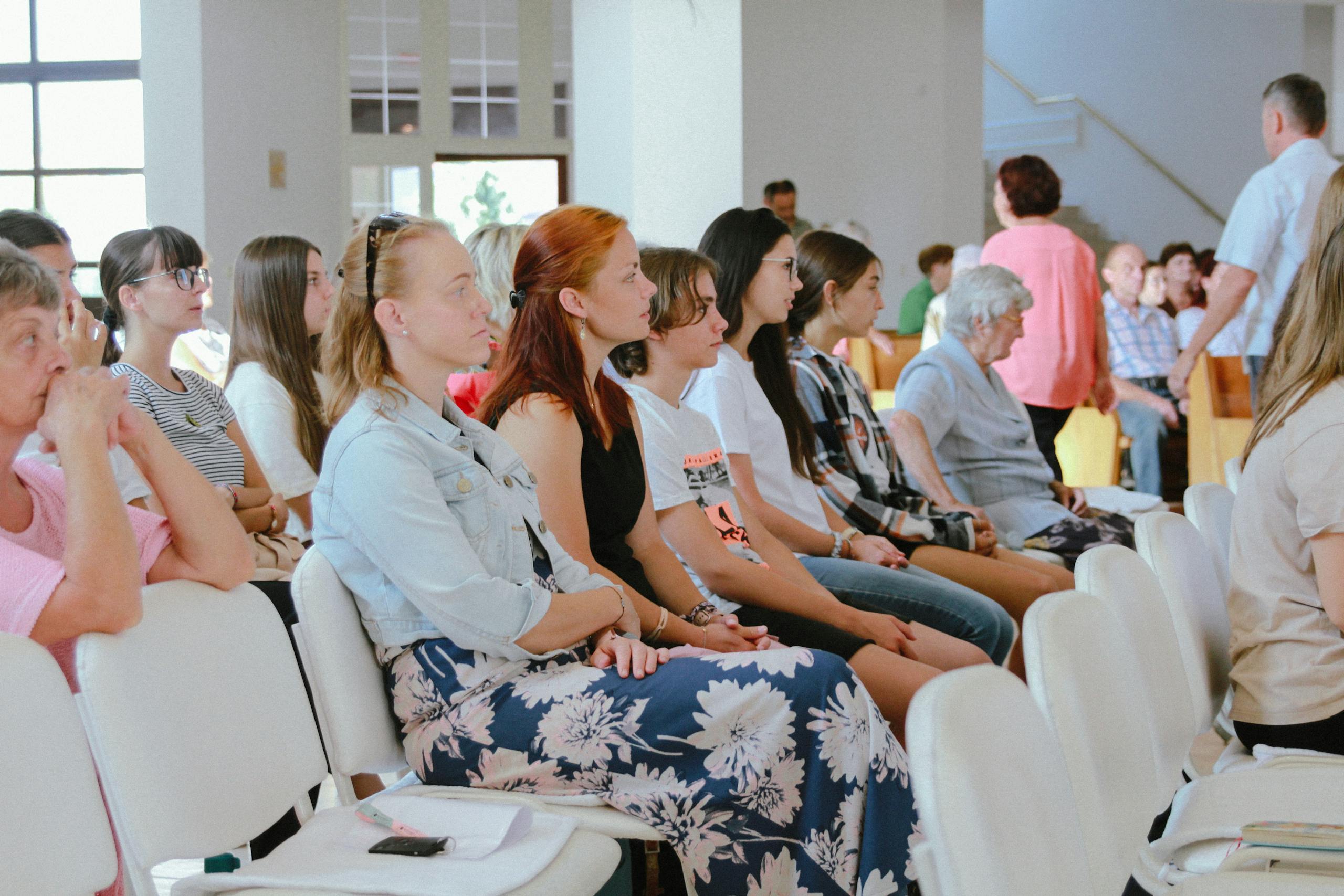 A diverse group of people sitting attentively during an indoor meeting or event, captured in a spacious room.